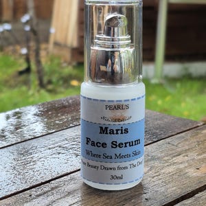 May include: A white and blue Maris Face Serum bottle with a silver pump dispenser. The label reads "Where Sea Meets Skin" and "30ml". The bottle is on a wooden surface.