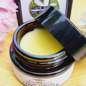 Könnte beinhalten: Ein kleines Glasgefäß mit schwarzem Deckel, das ein gelbes festes Parfum enthält. Das Gefäß hat ein weißes Etikett mit dem Text "PEARL'S Natural Cosmetic SOLID PERFUME TIARE GARDEN".