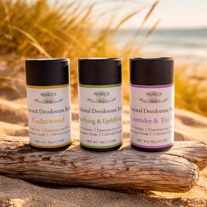 Könnte beinhalten: Drei natürliche Deodorant-Sticks mit schwarzen Deckeln und Etiketten. Die Etiketten lauten "Cedarwood", "Purifying & Uplifting" und "Lavender & Tea Tree". Jeder Stick wiegt 90g. Die Sticks liegen auf einem Stück Treibholz.