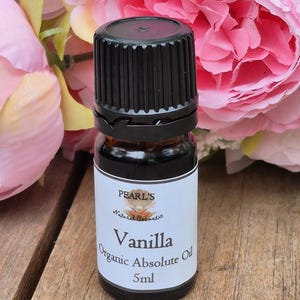 Può includere: Una piccola bottiglia di vetro da 5 ml di Olio Assoluto Biologico alla Vaniglia Pearl's. La bottiglia ha un tappo nero e un'etichetta bianca. Lo sfondo presenta fiori rosa e una superficie in legno.