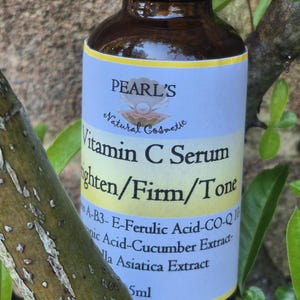May include: A brown glass bottle of Pearl's Vitamin C Serum. The label is light blue and yellow with text that reads "Vitamin C Serum Brighten/Firm/Tone". The bottle is surrounded by green leaves.
