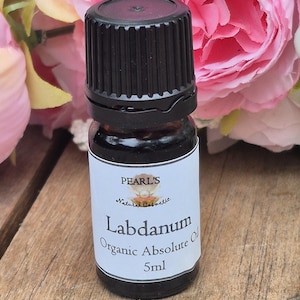 May include: A 5ml bottle of Labdanum Organic Absolute Oil with a black cap. The label reads "Pearl's Natural Cosmetic". The dark glass bottle sits on a wooden surface, with pink flowers in the background.