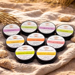 Könnte beinhalten: Eine Auswahl an natürlichen Deodorant-Balsamen in kleinen, runden, schwarzen Dosen. Jede Dose hat ein weißes Etikett mit dem Markennamen "PEARL'S Natural Cosmetic" und einem anderen Duftnamen. Die Balsame sind auf einer sandigen Oberfläche angeordnet.