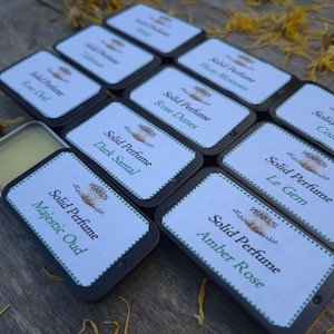 May include: A selection of small, rectangular solid perfume tins with white labels and black borders. The labels display the brand name "Pearls" and fragrance names such as "Rose Dunes" and "Le Gem". The background is a wooden surface.