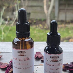 May include: Two brown glass bottles of facial serum with black droppers. The labels read "PEARL'S Natural Cosmetic Roseveil Elixir Facial Serum Radiance veiled in Rose". One bottle is 30ml, the other is 15ml. Dried rose petals are scattered around the bottles.