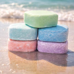 May include: A stack of six colorful, square-shaped bars of soap. The soaps are pastel shades of green, blue, pink, and purple. The soaps are stacked on a sandy surface with the ocean in the background.