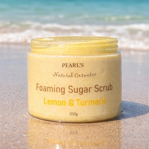 May include: A jar of Pearl's Foaming Sugar Scrub with Lemon & Turmeric. The yellow scrub is in a clear jar with a yellow lid. The label reads "Natural Cosmetic" and "200g". The jar is sitting on a sandy surface with the ocean in the background.