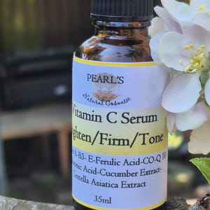 May include: A brown glass bottle of Pearl's Vitamin C Serum with a black cap. The label reads "Vitamin C Serum Brighten/Firm/Tone" and lists ingredients. The bottle is 35ml and is next to white flowers.