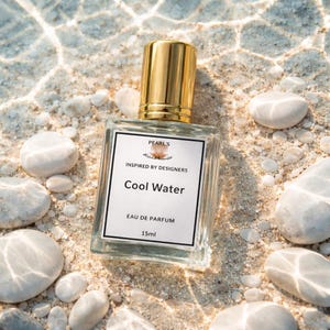 Op de afbeelding: Een doorzichtige glazen parfumfles met een gouden dop en een wit etiket. Het etiket bevat de tekst Pearl's, Inspired by Designers, Cool Water, Eau de Parfum en 15ml. De fles is omringd door gladde witte stenen op een zanderige ondergrond.