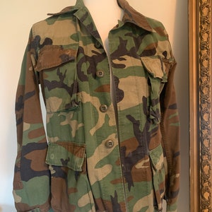 Könnte beinhalten: Eine grüne, braune und schwarze Tarnjacke mit Knopfleiste und zwei großen Taschen auf der Vorderseite.