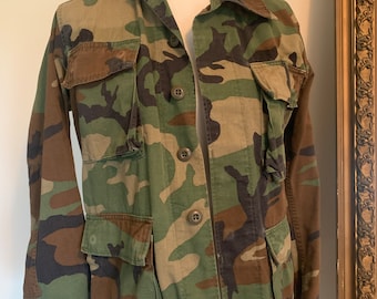May include: A green, brown, and black camouflage jacket with a button-up front and two large pockets on the front.