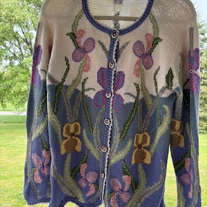 Könnte beinhalten: Cremefarbene Strickjacke mit einem floralen Iris-Design. Der Pullover hat eine Knopfleiste, einen blauen, gewellten Saum und einen runden Ausschnitt. Die Irisblüten sind lila, gelb und rosa, mit grünen Stielen und Blättern. Der Markenname "MAKIWAN" ist sichtbar.