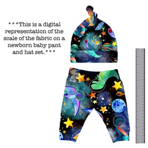 May include: A newborn baby pant and hat set with a space-themed print. The fabric features a dark blue background with stars, planets, and a spaceship. The pants are black with a blue, green, and yellow pattern. The hat is blue with a white knot on top.
