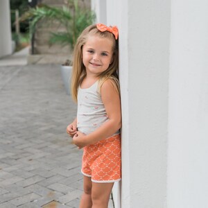 May include: A young girl wearing a white tank top with a floral print and orange shorts with a white pattern stands next to a white wall. She is wearing a pink bow in her hair and pink shoes.