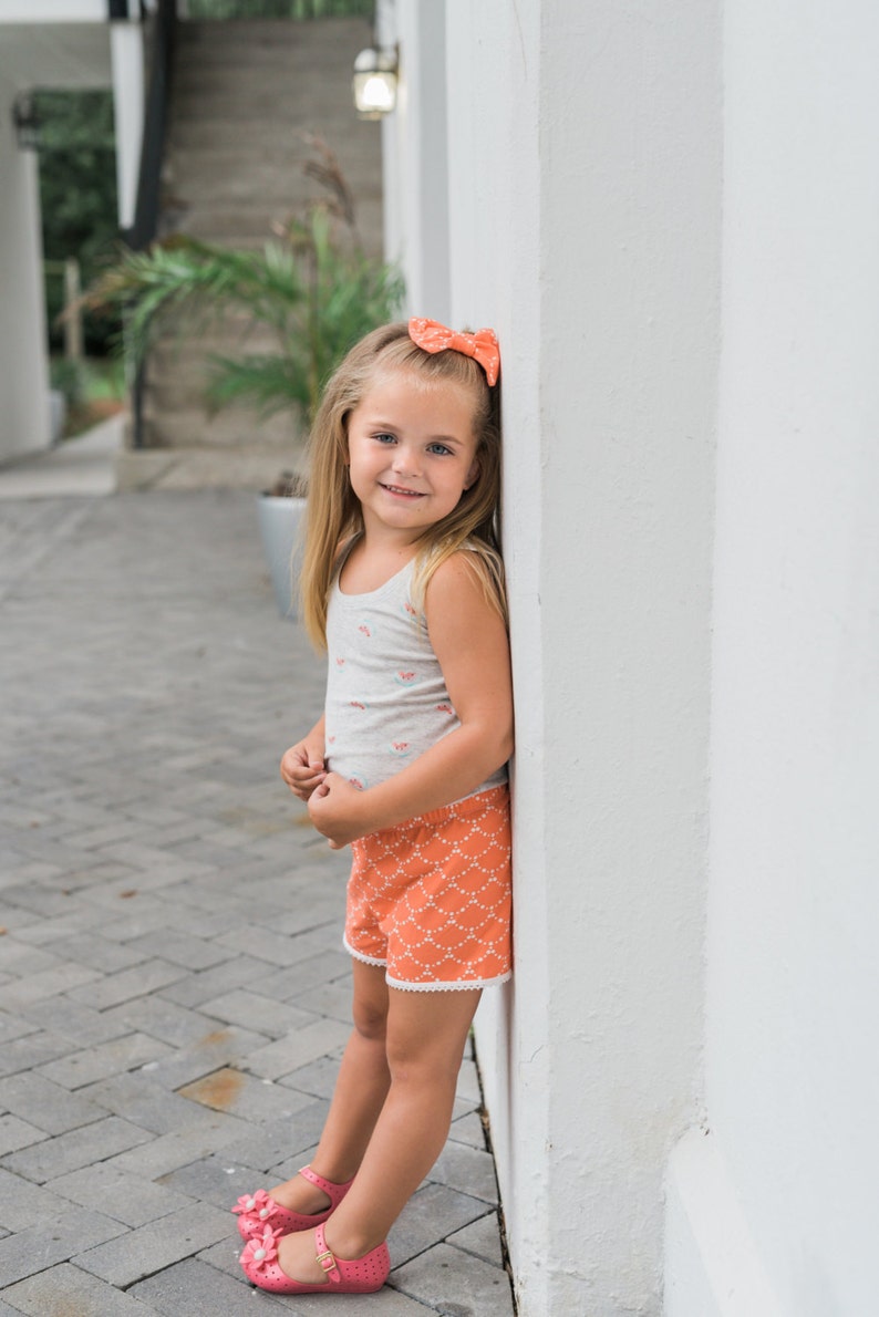 May include: A young girl wearing a white tank top with a floral print and orange shorts with a white pattern stands next to a white wall. She is wearing a pink bow in her hair and pink shoes.