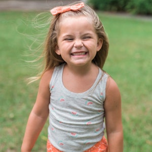 May include: A young girl with blonde hair wearing a grey tank top with watermelon print and orange shorts with a white pattern. She is smiling and has a pink bow in her hair.