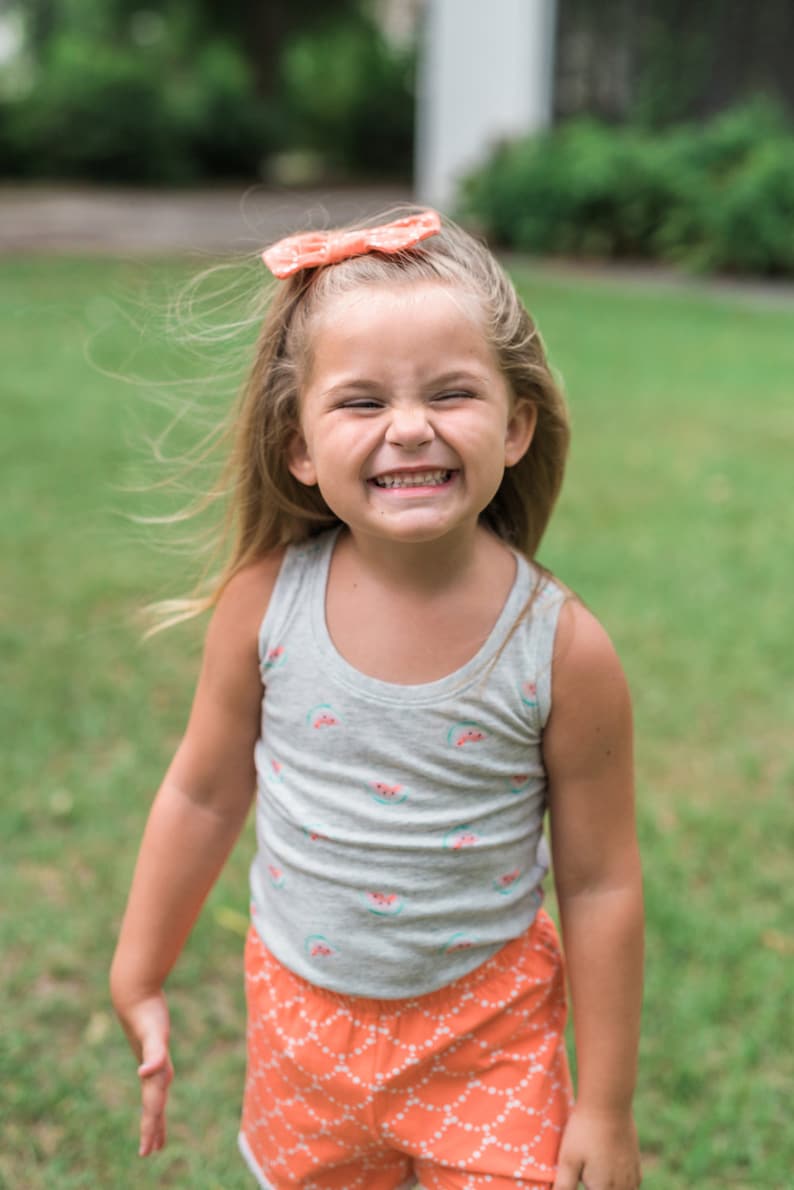 May include: A young girl with blonde hair wearing a grey tank top with watermelon print and orange shorts with a white pattern. She is smiling and has a pink bow in her hair.