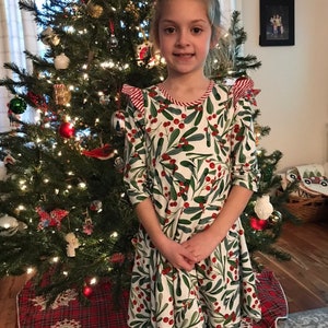 May include: A young girl wearing a white dress with a green and red holly berry pattern. She is standing in front of a Christmas tree decorated with red ornaments and lights.