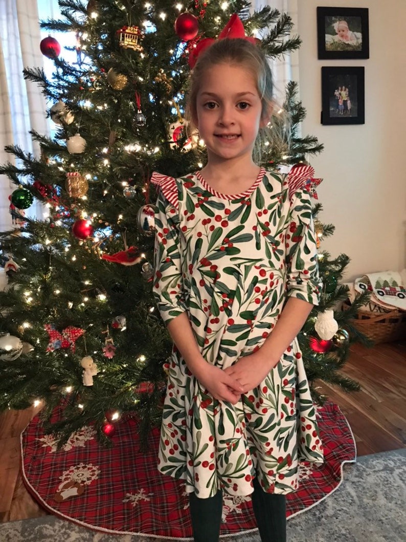 May include: A young girl wearing a white dress with a green and red holly berry pattern. She is standing in front of a Christmas tree decorated with red ornaments and lights.