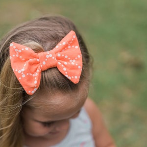May include: A young girl with long blonde hair wearing a coral bow with white polka dots.