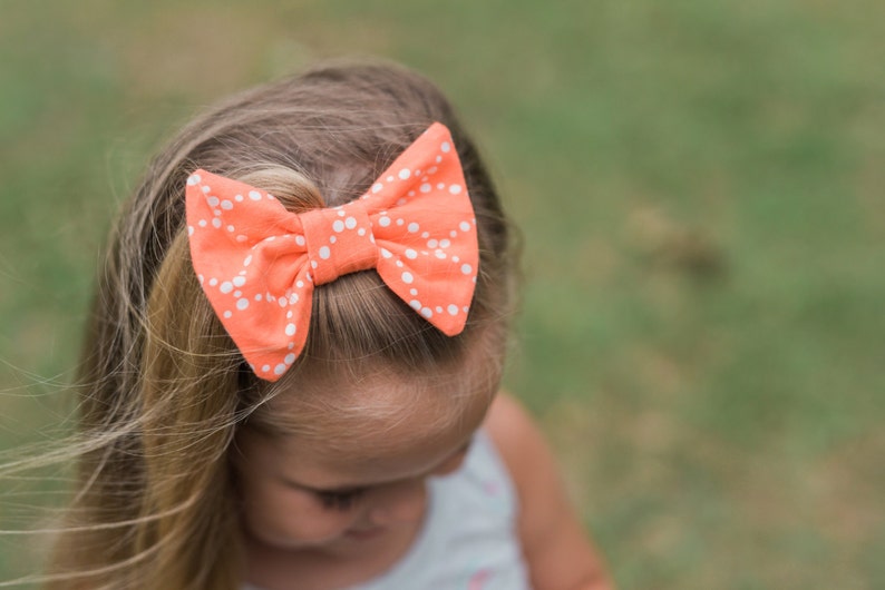 May include: A young girl with long blonde hair wearing a coral bow with white polka dots.