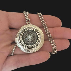 May include: Silver locket necklace with a wolf head design. The locket has an intricate design with a black background and a silver wolf head in the center.