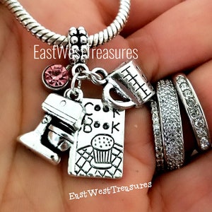 May include: Silver charm bracelet with a pink crystal charm, a measuring cup charm, a cookbook charm, and a stand mixer charm. The charms are attached to a silver chain bracelet. The bracelet is labeled "EastWestTreasures".