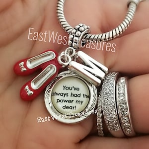 May include: Silver charm bracelet with a red ruby slipper charm, a silver letter "M" charm, and a round charm with the text "You've always had the power my dear!"