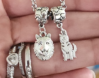 May include: Silver necklace with two charms, one of a wolf's head and the other of a dog. The charms are attached to a silver chain with paw print beads.