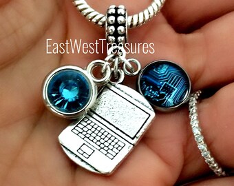 May include: Silver charm bracelet with a laptop charm, a blue crystal charm, and a charm with a blue circuit board design. The charm bracelet is on a hand.