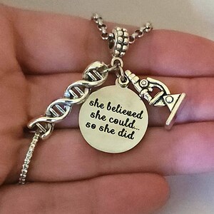 May include: Silver necklace with a charm that says "she believed she could... so she did". The charm is attached to a silver DNA strand and a silver microscope charm.