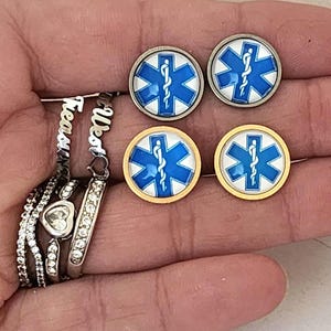 May include: A pair of earrings with a blue and white medical symbol on a gold and silver background. The earrings are round and have a shiny finish.