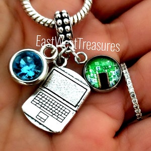 May include: A silver charm bracelet with a laptop charm, a blue crystal charm, and a charm with a green circuit board design. The charm bracelet is on a hand.