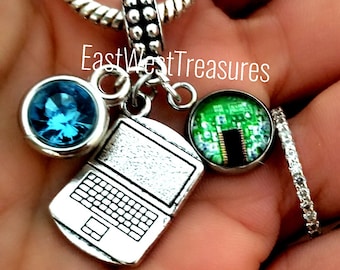 May include: A silver charm bracelet with a laptop charm, a blue crystal charm, and a charm with a green circuit board design. The charm bracelet is on a hand.