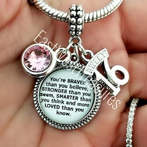 May include: Silver charm bracelet with a pendant featuring a pink gemstone, a 'Sweet 16' charm, and a round charm with the text "You're BRAVER than you believe..." A ring with small clear stones is also visible.