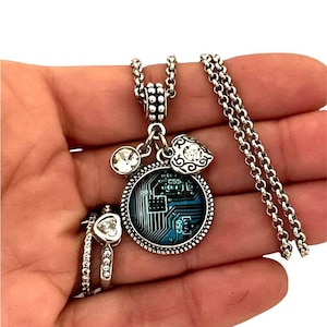 May include: A silver chain necklace with a charm featuring a blue and black circuit board design. The charm is surrounded by a silver frame and has a small silver heart and crystal charm attached.