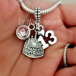 May include: Silver charm bracelet with a pink crystal, a cake charm with the text "Happy Birthday", and a charm with the number "13".