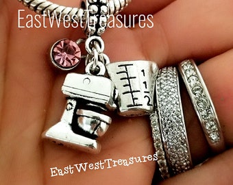 May include: Silver charm bracelet with a pink crystal charm, a silver measuring cup charm, and a silver stand mixer charm. The charm bracelet is on a hand wearing several silver rings.