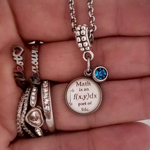 May include: Silver-toned necklace with a round pendant that reads "Math is an integral part of life." A small blue gemstone hangs below the pendant. Several rings with the words "West" and "Treasure" are also visible.