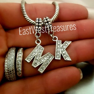 May include: Silver charm bracelet with two letter charms, one with the letter M and one with the letter K, both adorned with clear rhinestones. The charms are attached to a silver snake chain bracelet.