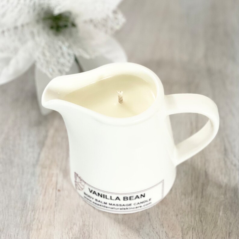 May include: White ceramic pitcher candle with a vanilla bean scent. The candle is filled with white wax and has a single wick. The label on the front reads "Vanilla Bean Body Balm Massage Candle".