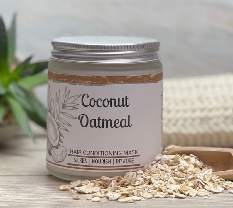 May include: A glass jar with a silver lid labeled "Coconut Oatmeal Hair Conditioning Mask." The label features a coconut and palm frond illustration. Rolled oats are scattered around the jar. The text on the label also says "Silken | Nourish | Restore."