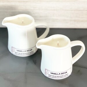 May include: Two white ceramic pitcher candles with a vanilla bean scent. The candles have a label that reads "Vanilla Bean Body Balm Massage Candle" and a website address.