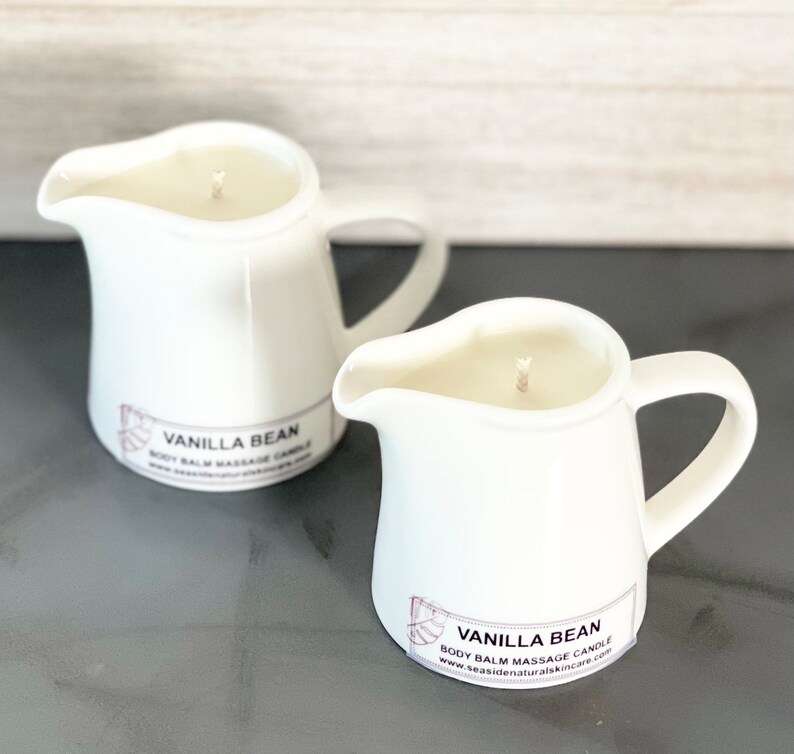 May include: Two white ceramic pitcher candles with a vanilla bean scent. The candles have a label that reads "Vanilla Bean Body Balm Massage Candle" and a website address.