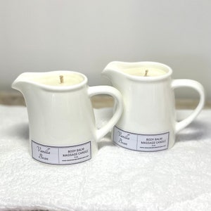 May include: Two white ceramic pitcher candles with a vanilla bean scent. The candles are labelled "Vanilla Bean" and "Body Balm Massage Candle".
