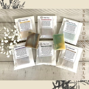 May include: Six bars of handmade soap wrapped in white paper bags with labels. The labels have nautical themed logos and list the scents: Lemon Peel, Wild Rose, Seas the Day, Calming Lavender, Man Cave, and Cedarwood. The soaps are arranged on a light wood surface.