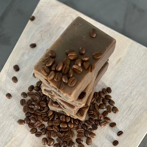May include: A stack of three brown coffee soap bars with coffee beans scattered around them. The soap bars are rectangular and have a smooth, creamy texture.