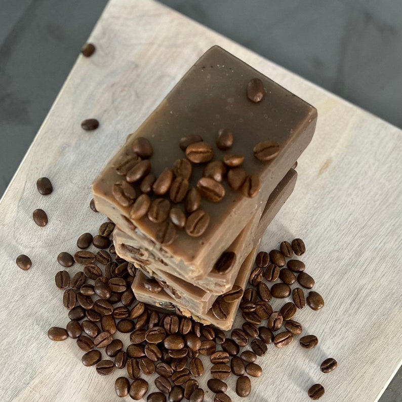 May include: A stack of three brown coffee soap bars with coffee beans scattered around them. The soap bars are rectangular and have a smooth, creamy texture.