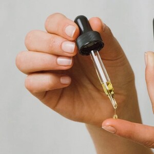 May include: A hand holding a clear glass dropper bottle with a black cap. A drop of liquid is falling from the dropper onto a finger.
