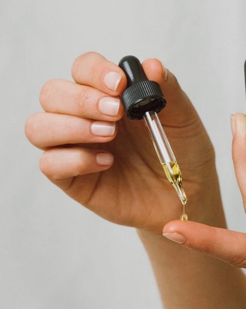 May include: A hand holding a clear glass dropper bottle with a black cap. A drop of liquid is falling from the dropper onto a finger.